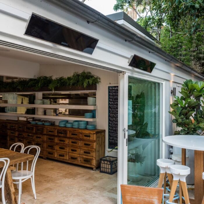 Modern café interior featuring a wooden dining table, white chairs, and open kitchen with shelves of plates and a chalkboard menu. Natural light flows through large glass doors, enhancing the inviting atmosphere.
