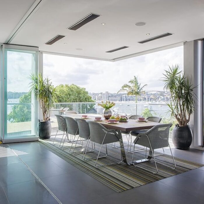 Bright and modern dining area with a large table and stylish chairs, featuring a view of the waterfront through glass walls, surrounded by greenery and decorative plants.