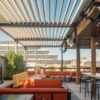 Rooftop dining area with modern decor, featuring orange seating, a fire pit, and a spacious layout under a slatted pergola, overlooking city buildings.