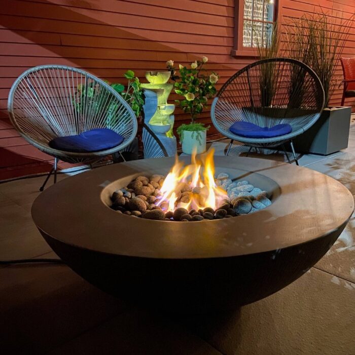 Cozy outdoor seating area featuring a modern fire pit surrounded by decorative stones, two stylish chairs with blue cushions, and lush plants, creating a warm and inviting atmosphere at night.
