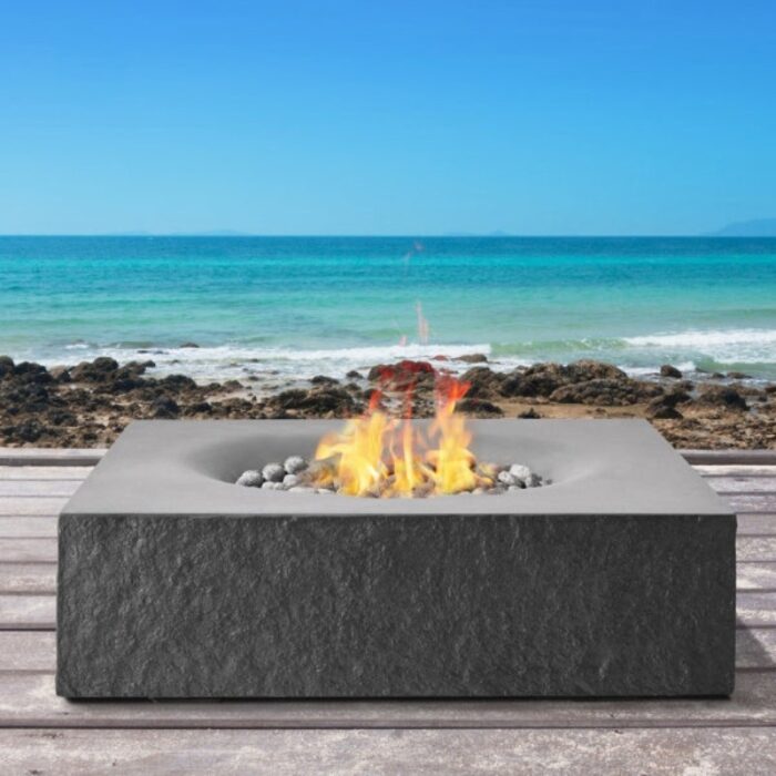 Modern outdoor fire pit on a wooden deck overlooking the ocean, featuring flames and decorative stones.