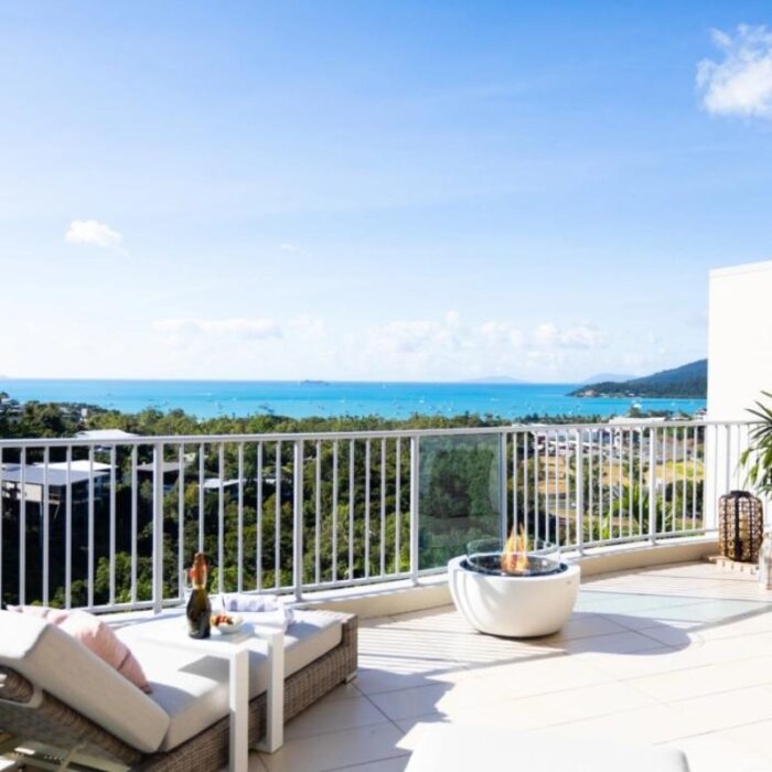 Modern balcony with a stunning ocean view, featuring a comfortable lounge chair, a small table with refreshments, and a stylish fire pit. Lush greenery and coastal landscape in the background under a clear blue sky.