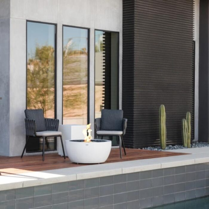 Modern outdoor seating area featuring two stylish chairs and a contemporary fire pit, set against a sleek architectural backdrop with large windows and cactus plants.