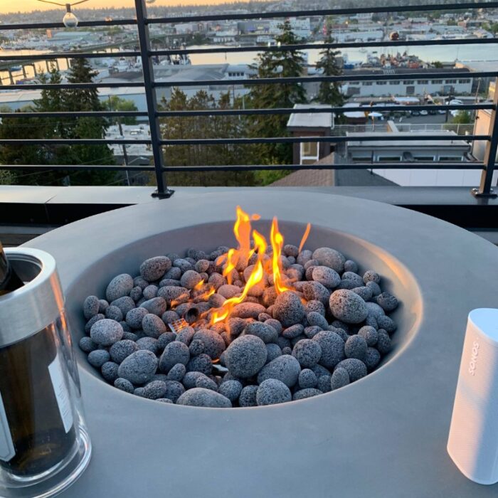 Modern rooftop fire pit with glowing flames and smooth black stones, overlooking a scenic urban landscape at sunset, accompanied by a wine bottle and a portable speaker.