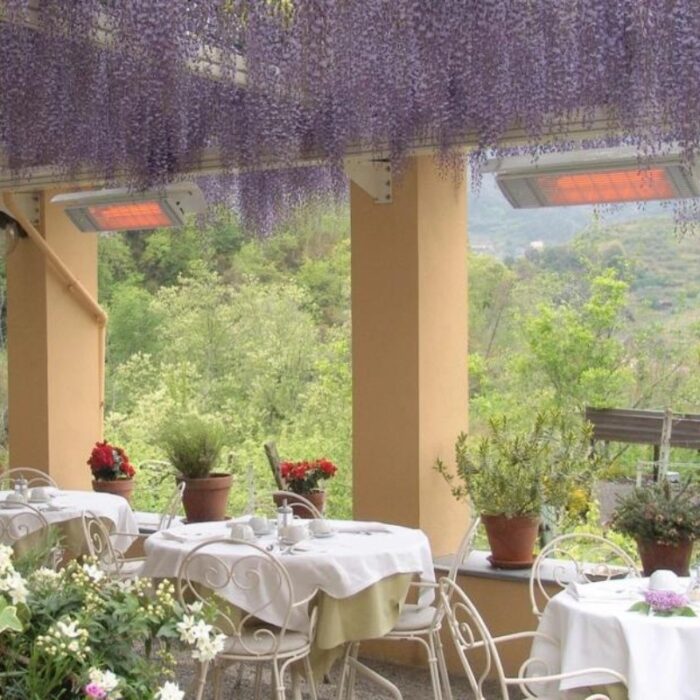Outdoor dining area with white tablecloths, surrounded by potted plants and blooming flowers, under a canopy of purple wisteria.