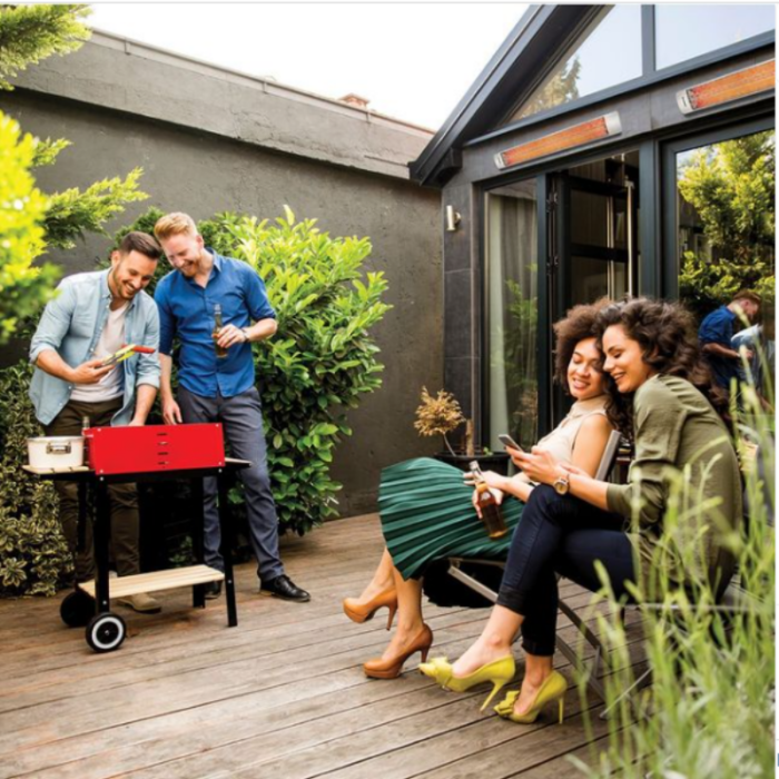 Group of friends enjoying a barbecue in a backyard, with two men preparing food on a red grill and two women relaxing on a bench, all surrounded by greenery.