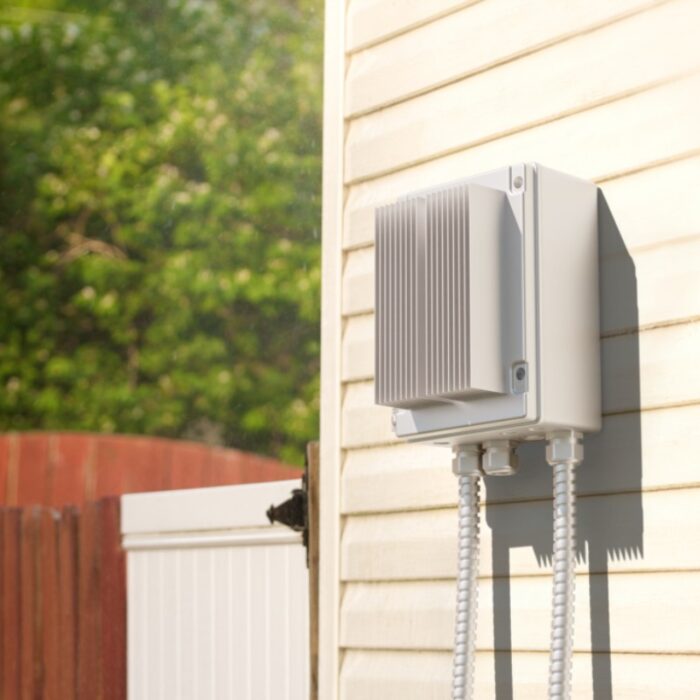 Outdoor electrical box mounted on a house wall with protective cables, surrounded by greenery.