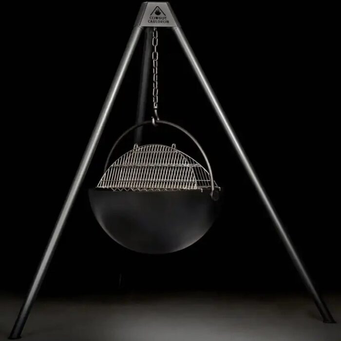 Hanging metal cauldron grill with a wire grate, suspended from a tripod stand against a dark background.