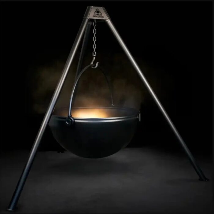 A black cooking cauldron suspended from a metallic tripod, emitting a soft glow and steam against a dark background.
