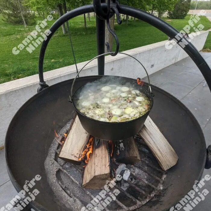 Cooking a hearty stew over an open fire in a cast iron pot, with visible smoke and wooden logs burning beneath it, surrounded by a green outdoor setting.