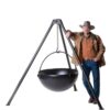 Man in cowboy attire leaning against a large black cooking cauldron on a tripod stand, emphasizing outdoor cooking and rugged lifestyle.