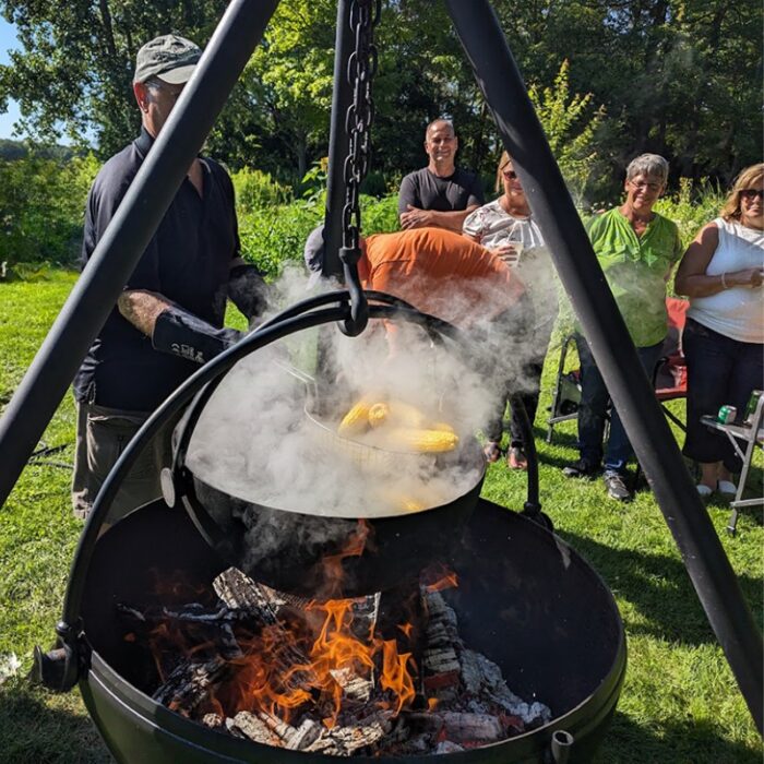 Outdoor cooking scene with people gathered around a fire, steaming corn in a hanging pot over an open flame.