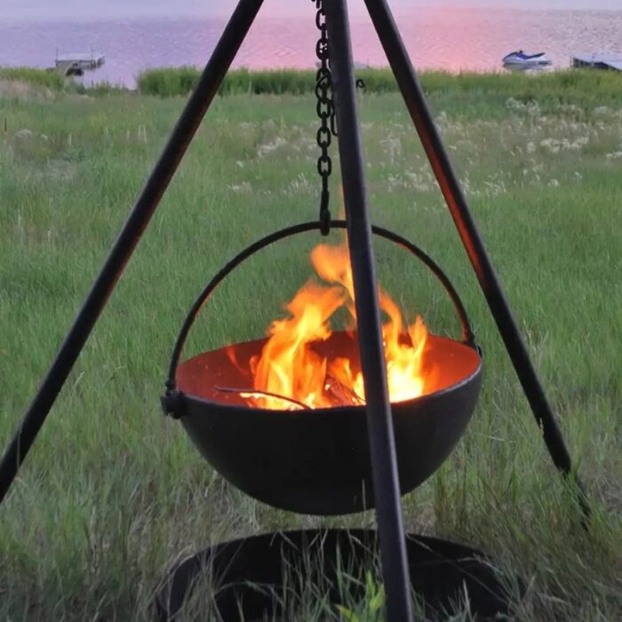 A black cauldron filled with flames hangs from a tripod over a grassy area, with a serene waterfront view in the background during sunset.