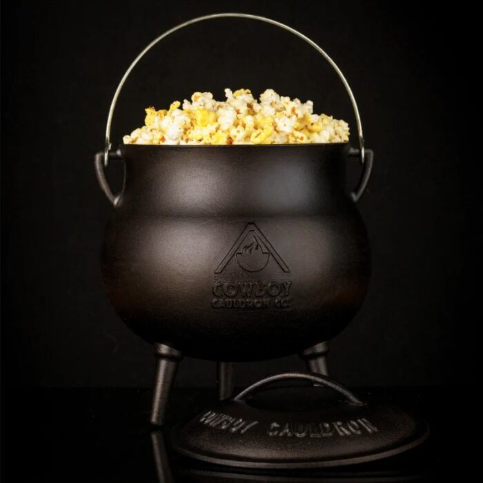 Black cast iron cauldron filled with popcorn, featuring a metal handle and standing on three legs, with the Cowboy Cauldron logo visible.