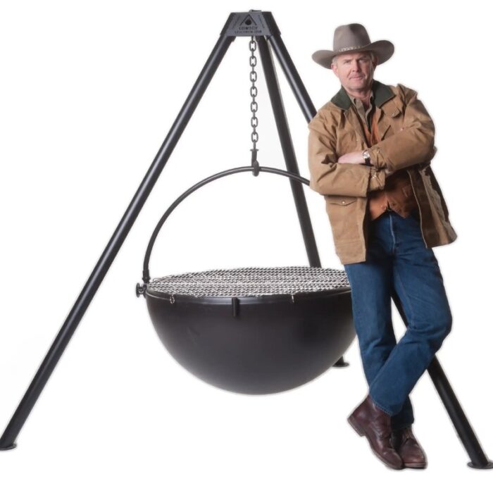 A cowboy leaning casually against a large, black cauldron-style grill, showcasing a sturdy tripod stand and chain support. He is dressed in a brown jacket, denim jeans, and a cowboy hat, exuding a rustic, outdoor cooking vibe.