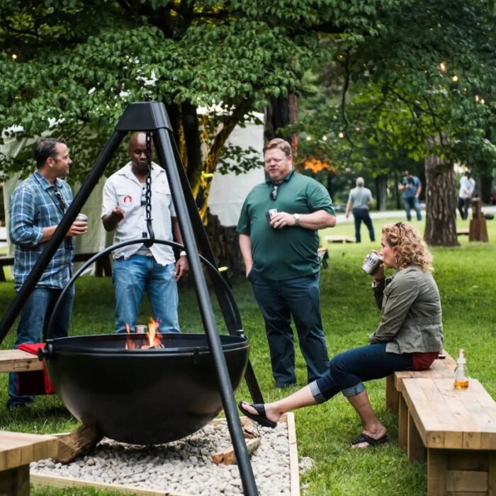 Group of friends enjoying a casual gathering around a fire pit in a park, with one person seated and sipping a drink while others engage in conversation.