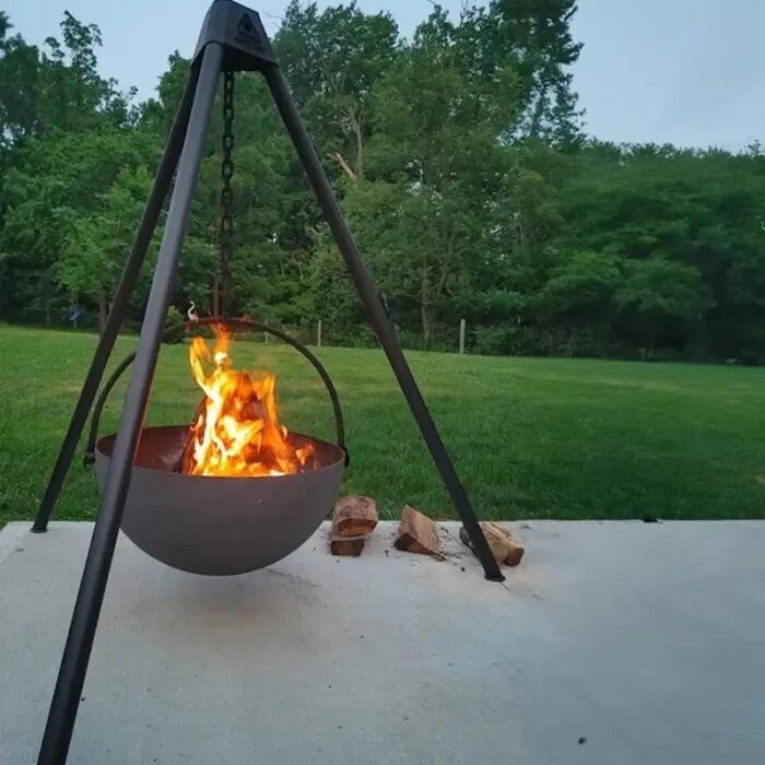 Hanging fire pit with flames, set on a concrete surface surrounded by grass and firewood, in a landscaped outdoor area.