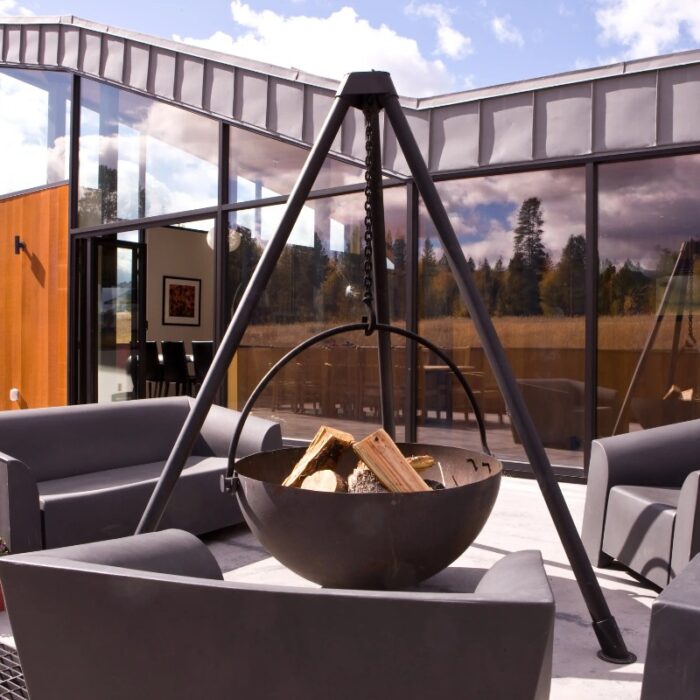 Modern outdoor seating area featuring a large hanging fire pit with logs, surrounded by sleek lounge chairs, set against a backdrop of glass walls and a scenic landscape.