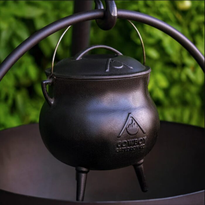 Cast iron pot hanging on a metal stand with greenery in the background, ideal for outdoor cooking and camping.
