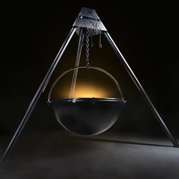 A large black cooking cauldron suspended from a metal A-frame, illuminated against a dark background, showcasing its reflective surface and structural details.