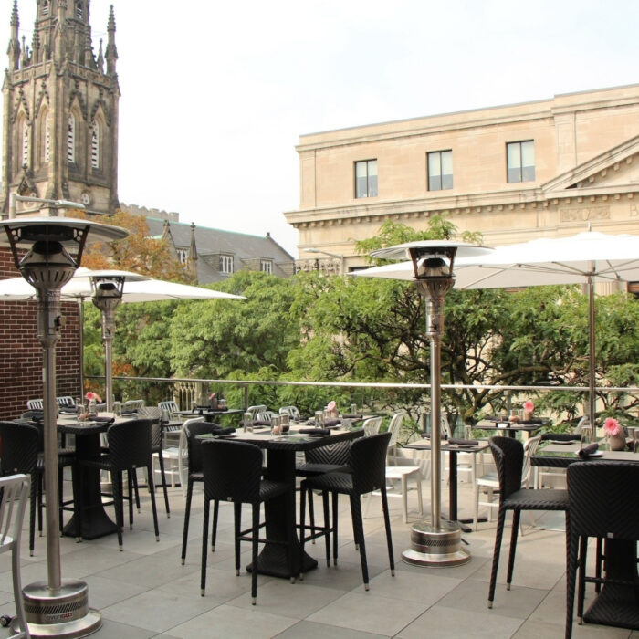Outdoor dining area with tables and chairs, featuring patio heaters and umbrellas, set against a backdrop of historic buildings and greenery.