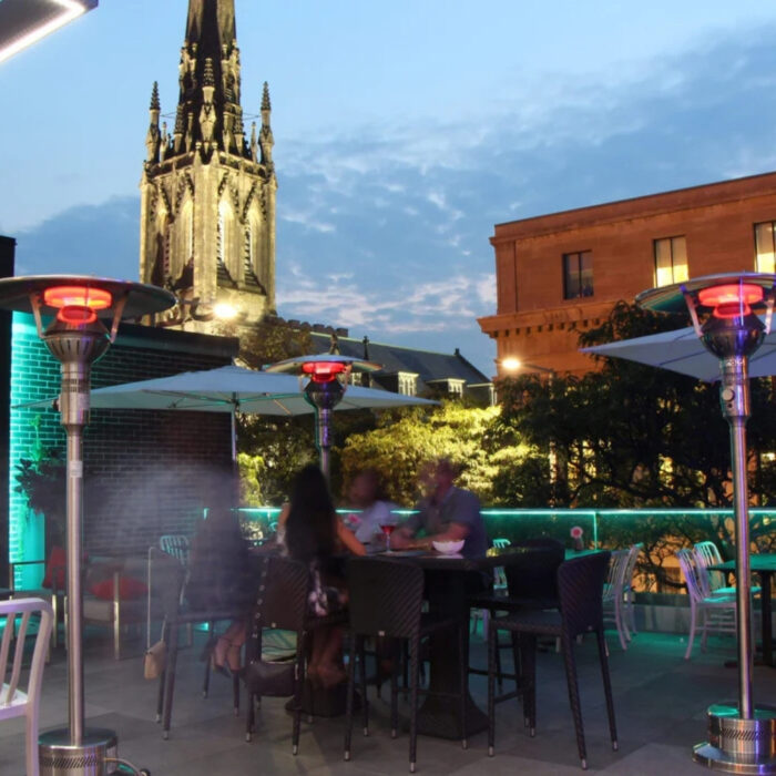 Rooftop dining area with patio heaters, featuring diners enjoying the evening, against a backdrop of a lit church steeple and city buildings during twilight.
