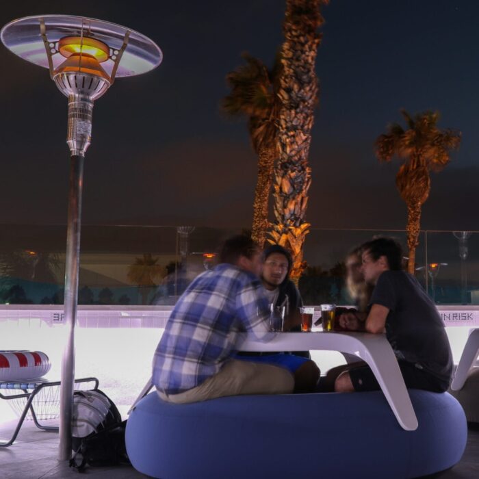 Group of friends socializing around a modern table with drinks at a vibrant outdoor setting, illuminated by a patio heater and surrounded by palm trees at night.
