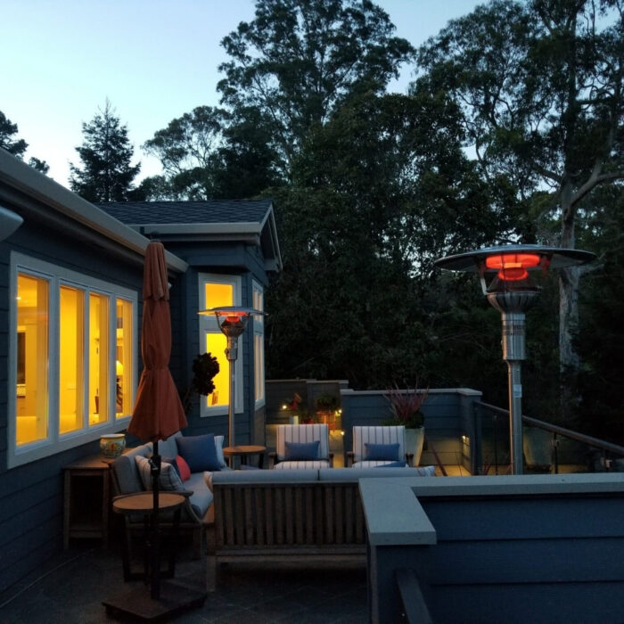 Cozy outdoor patio with seating, a large umbrella, and patio heaters, illuminated by warm light from the house and surrounded by trees at dusk.