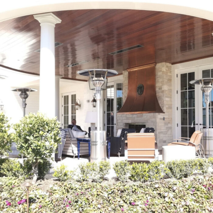 Outdoor seating area with comfortable furniture, a stone fireplace, and patio heaters, surrounded by neatly trimmed shrubs and colorful flowers.