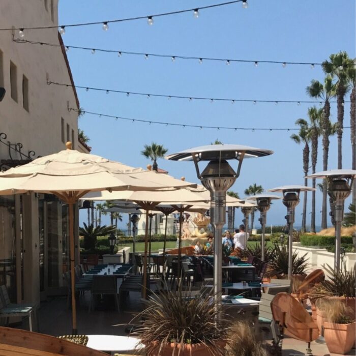 Outdoor dining area with patio umbrellas, heating lamps, and palm trees, overlooking the ocean on a sunny day.