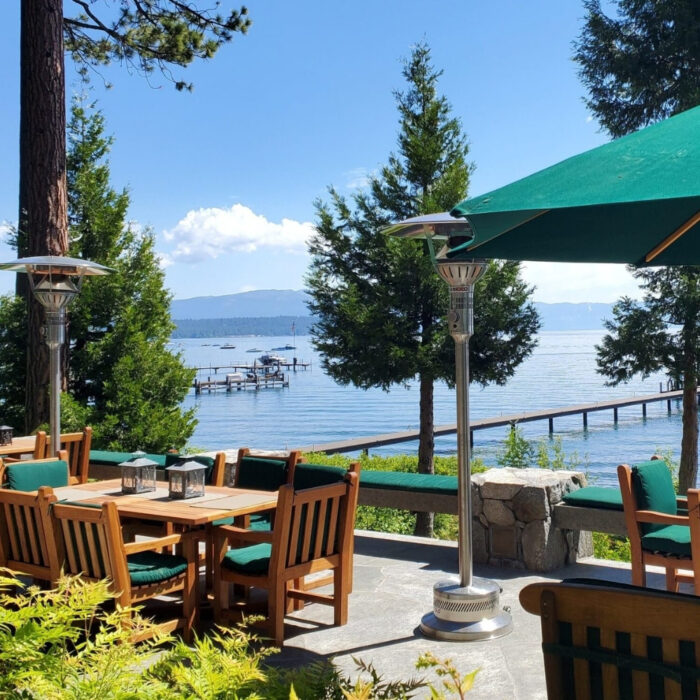 Scenic lakeside dining area with wooden tables and chairs, surrounded by greenery, overlooking a calm lake and a dock with boats.