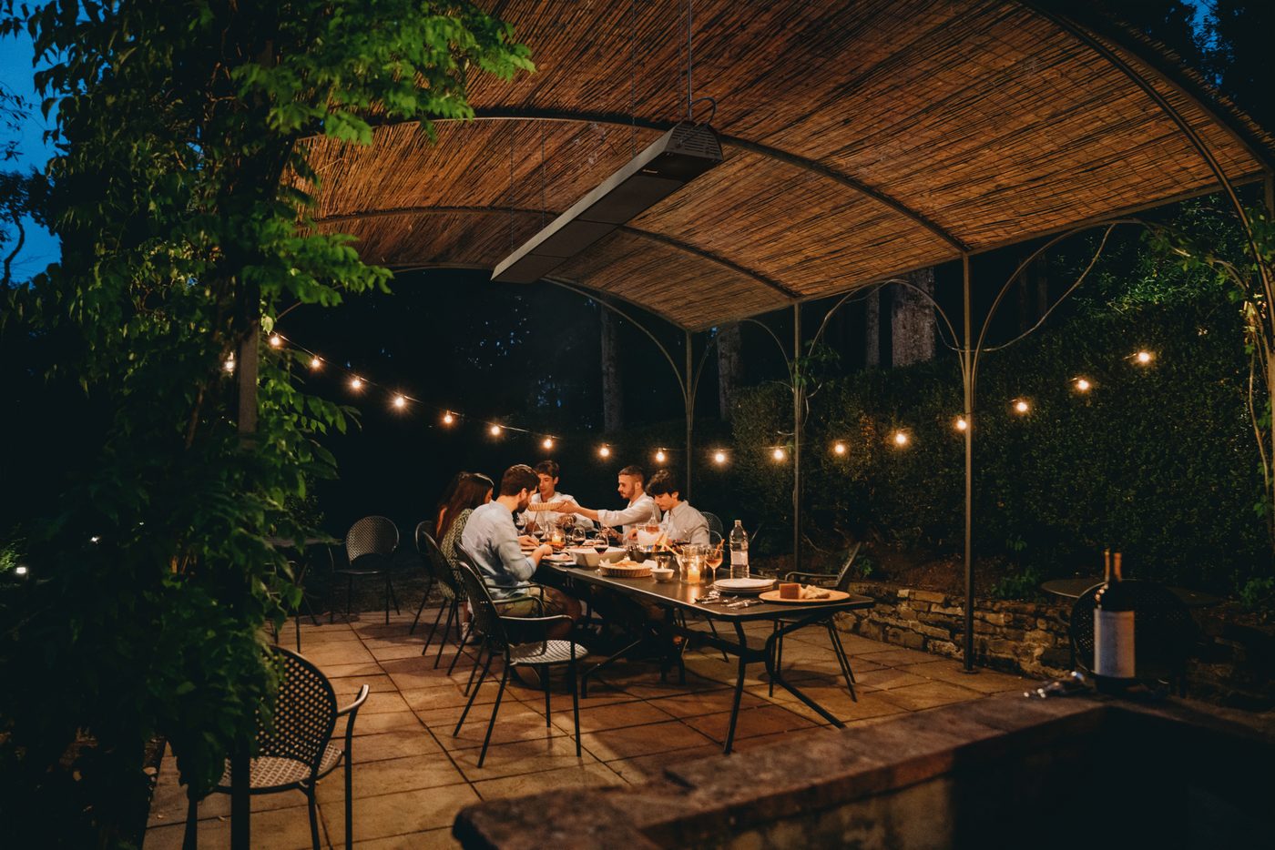 Diners at a long table on a covered patio warmed by an overhead infrared tube heater at night